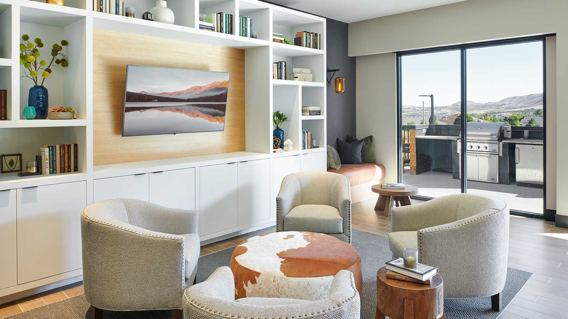 Epoque Golden resident lounge with armchairs, built-in shelving, wall-mounted TV and doors opening to an outdoor grilling terrace.