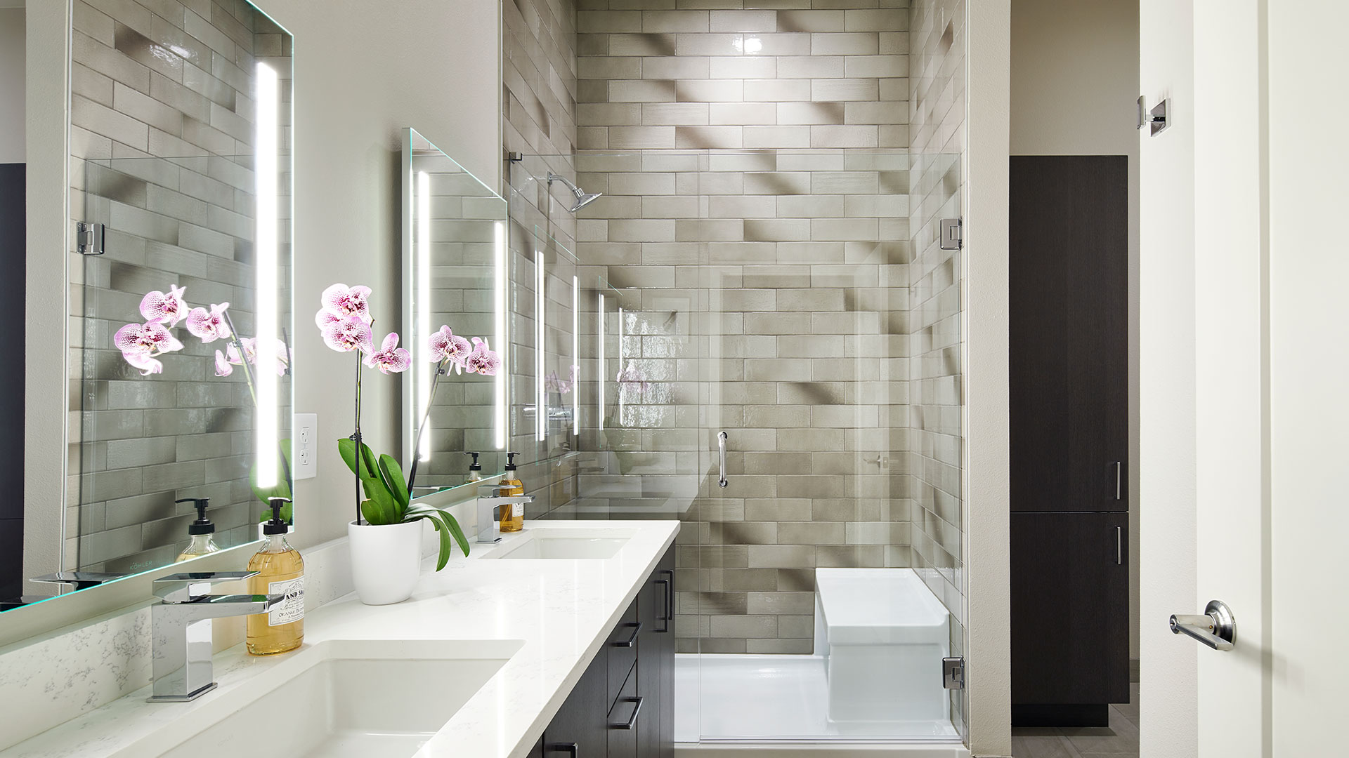Epoque Golden apartment bathroom with glass-enclosed tiled shower, double vanity, illuminated mirrors and modern fixtures.