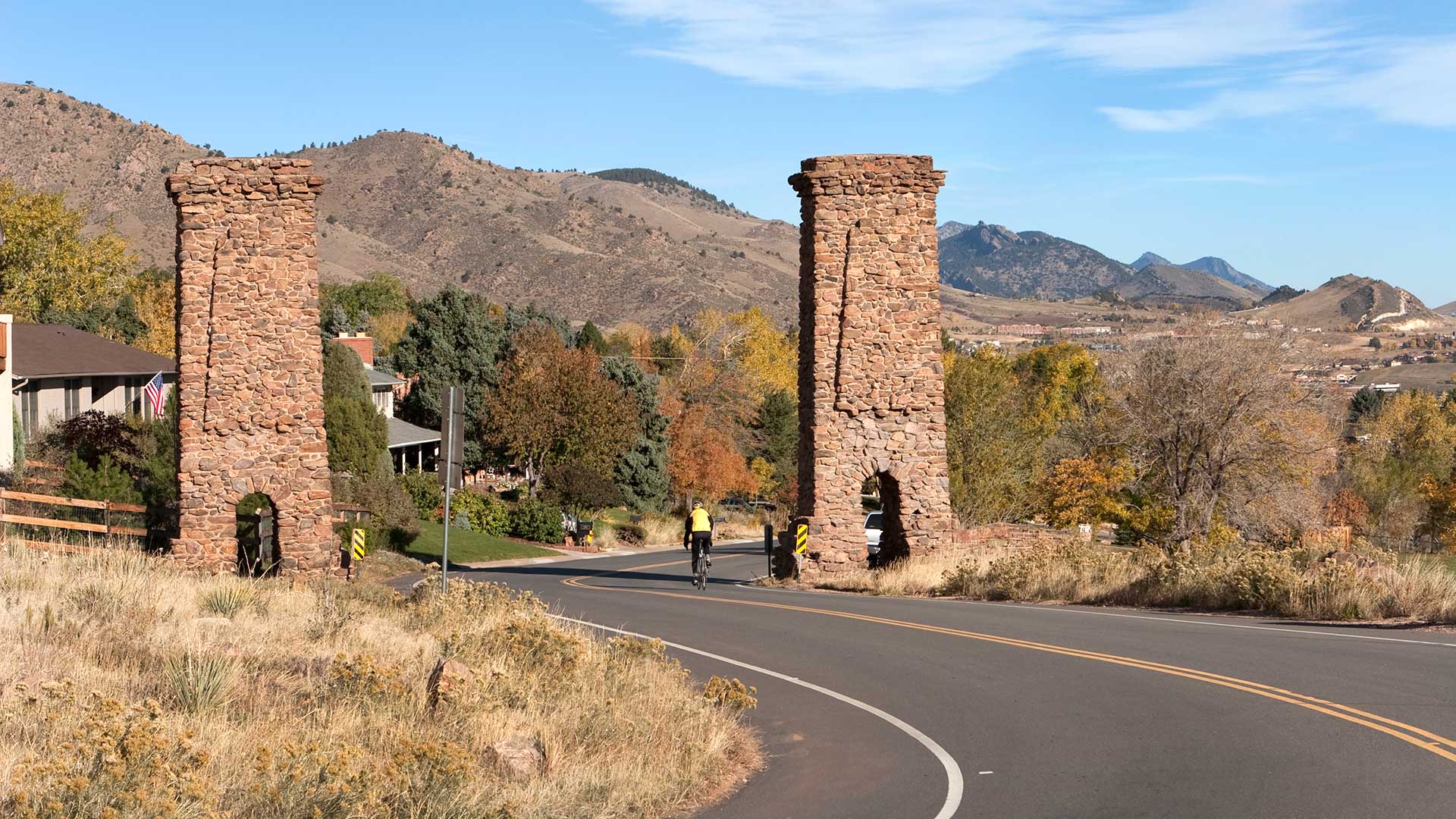 Cyclist riding through Golden, Colorado, between historic stone pillars with foothills and autumn trees in the background.